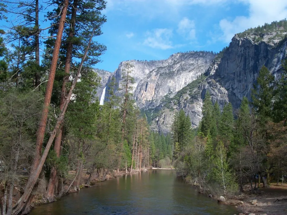 A wide river flows through a forest towards a tall waterfall cascading down a rocky mountain.