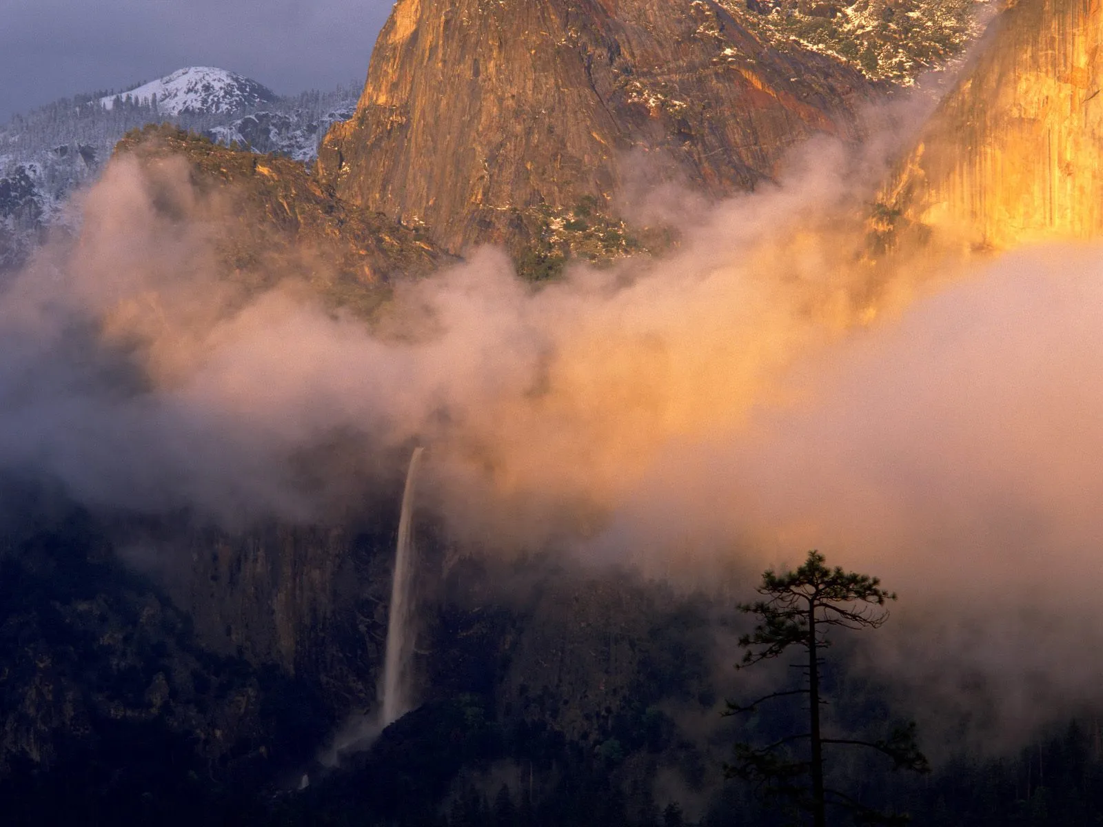 A tall waterfall cascades down a rocky cliff face, partially obscured by soft, pinkish clouds at sunrise.