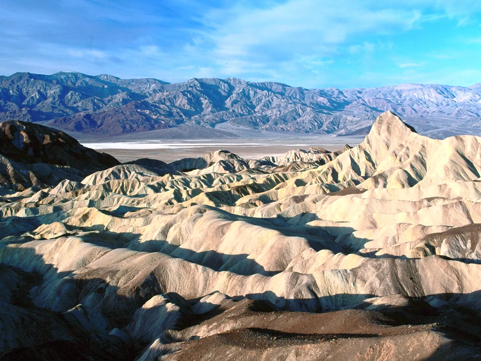 A vast desert landscape with eroded hills and distant mountains under a bright blue sky.