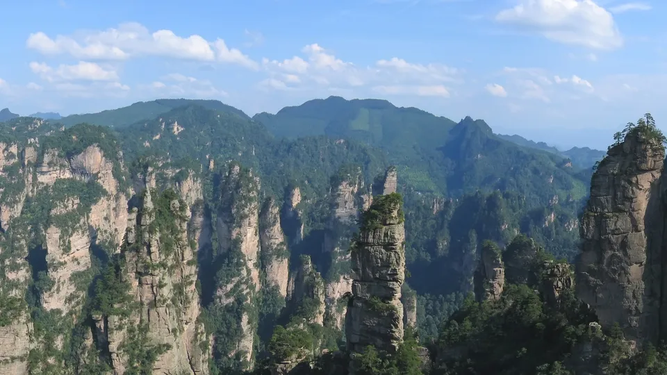 A panoramic view of numerous tall, slender sandstone pillars covered in green vegetation under a blue sky with scattered clouds.
